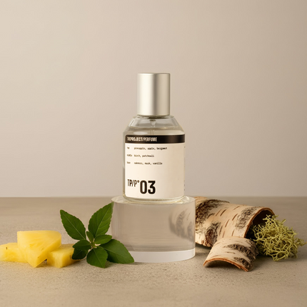 A clear glass perfume bottle with a silver cap and a white label reading 'TPP 03' stands on a clear acrylic riser. The bottle is styled on a beige surface with pineapple slices, mint leaves, birch bark, and dried moss against a neutral beige background.