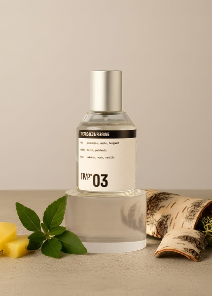 A clear glass perfume bottle with a silver cap and a white label reading 'TPP 03' stands on a clear acrylic riser. The bottle is styled on a beige surface with pineapple slices, mint leaves, birch bark, and dried moss against a neutral beige background.