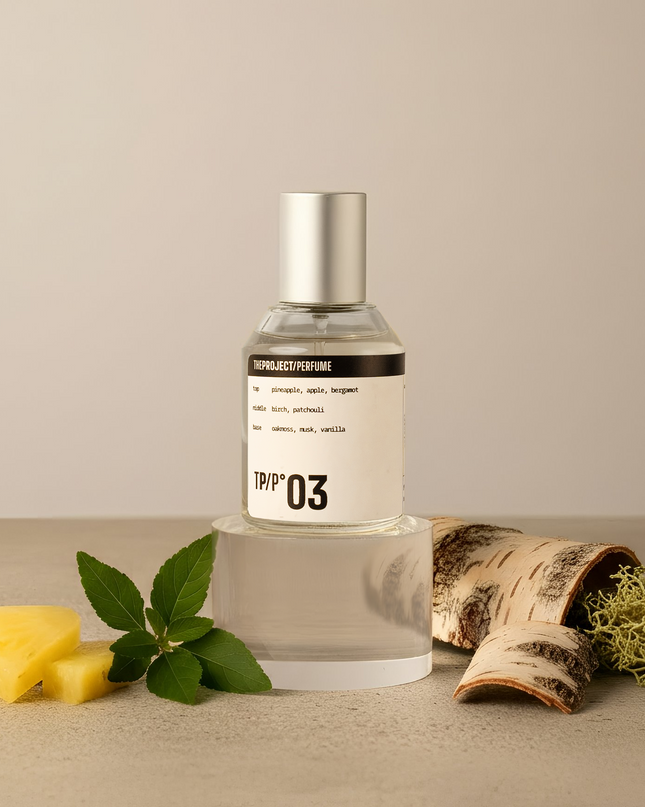 A clear glass perfume bottle with a silver cap and a white label reading 'TPP 03' stands on a clear acrylic riser. The bottle is styled on a beige surface with pineapple slices, mint leaves, birch bark, and dried moss against a neutral beige background.